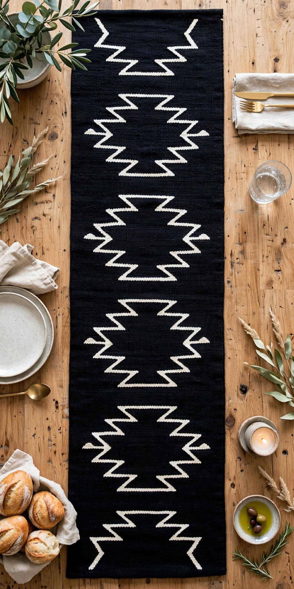 A long black woven table runner with a white geometric zigzag pattern, laid flat on a rustic wooden table surrounded by dining props like plates, bread, candles, and greenery.