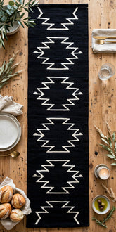 A long black woven table runner with a white geometric zigzag pattern, laid flat on a rustic wooden table surrounded by dining props like plates, bread, candles, and greenery.