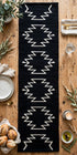 A long black woven table runner with a white geometric zigzag pattern, laid flat on a rustic wooden table surrounded by dining props like plates, bread, candles, and greenery.