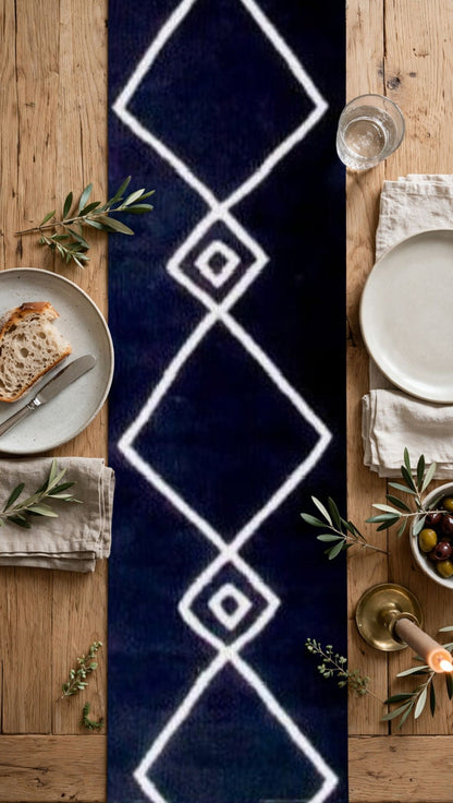 A long runner rug laid down on a wooden dining table, featuring a bold white geometric diamond pattern on a dark navy blue background. The table is styled with plates, napkins, olive branches, and a glass of water.