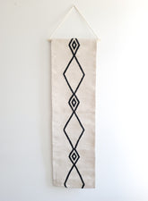 A vertical, handwoven wall hanging tapestry with a geometric diamond pattern in black on a cream background, with the top part folded over for display.