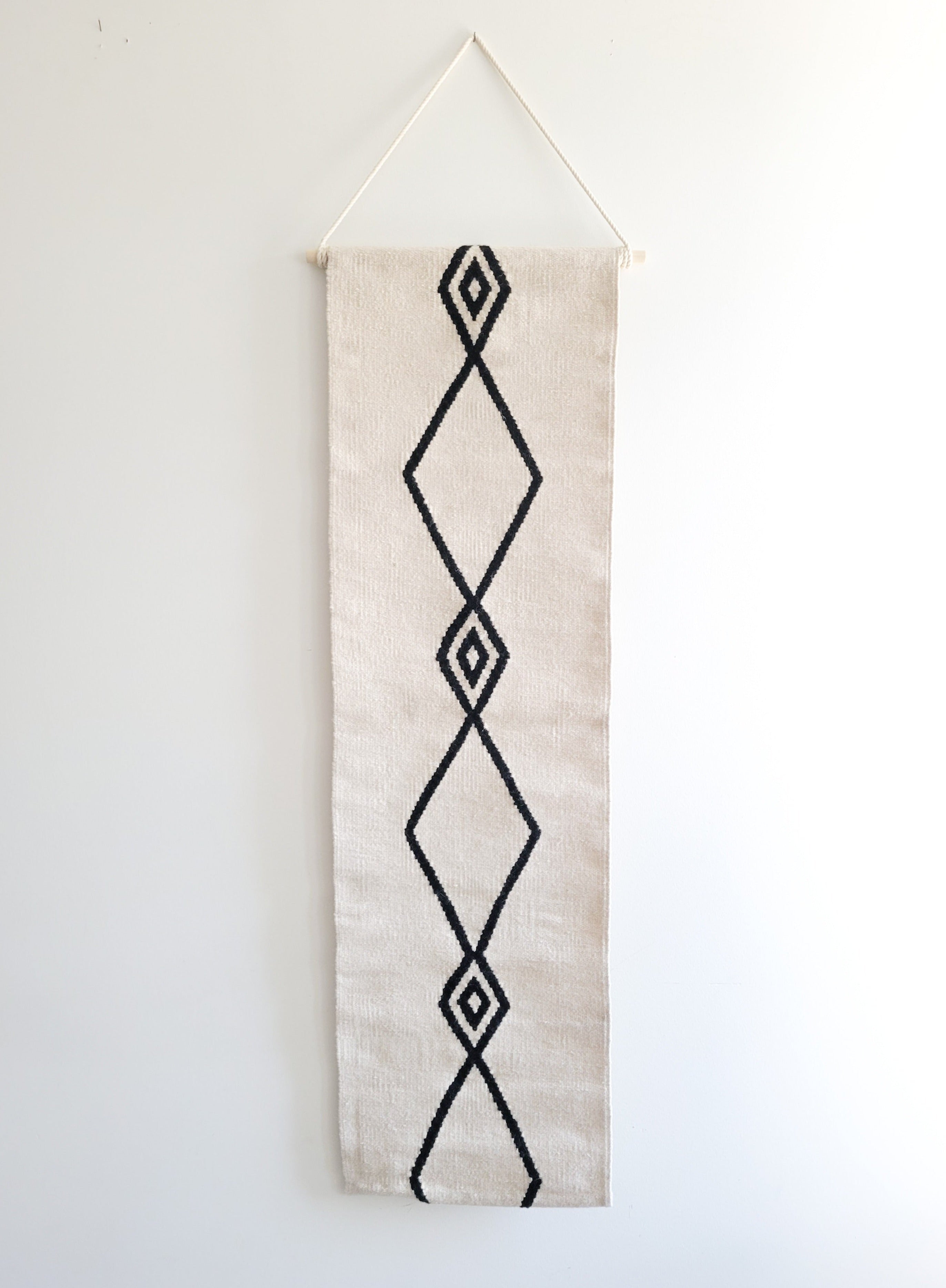 A vertical, handwoven wall hanging tapestry with a geometric diamond pattern in black on a cream background, with the top part folded over for display.