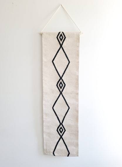 A vertical, handwoven wall hanging tapestry with a geometric diamond pattern in black on a cream background, with the top part folded over for display.