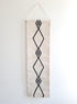 A vertical, handwoven wall hanging tapestry with a geometric diamond pattern in black on a cream background, with the top part folded over for display.