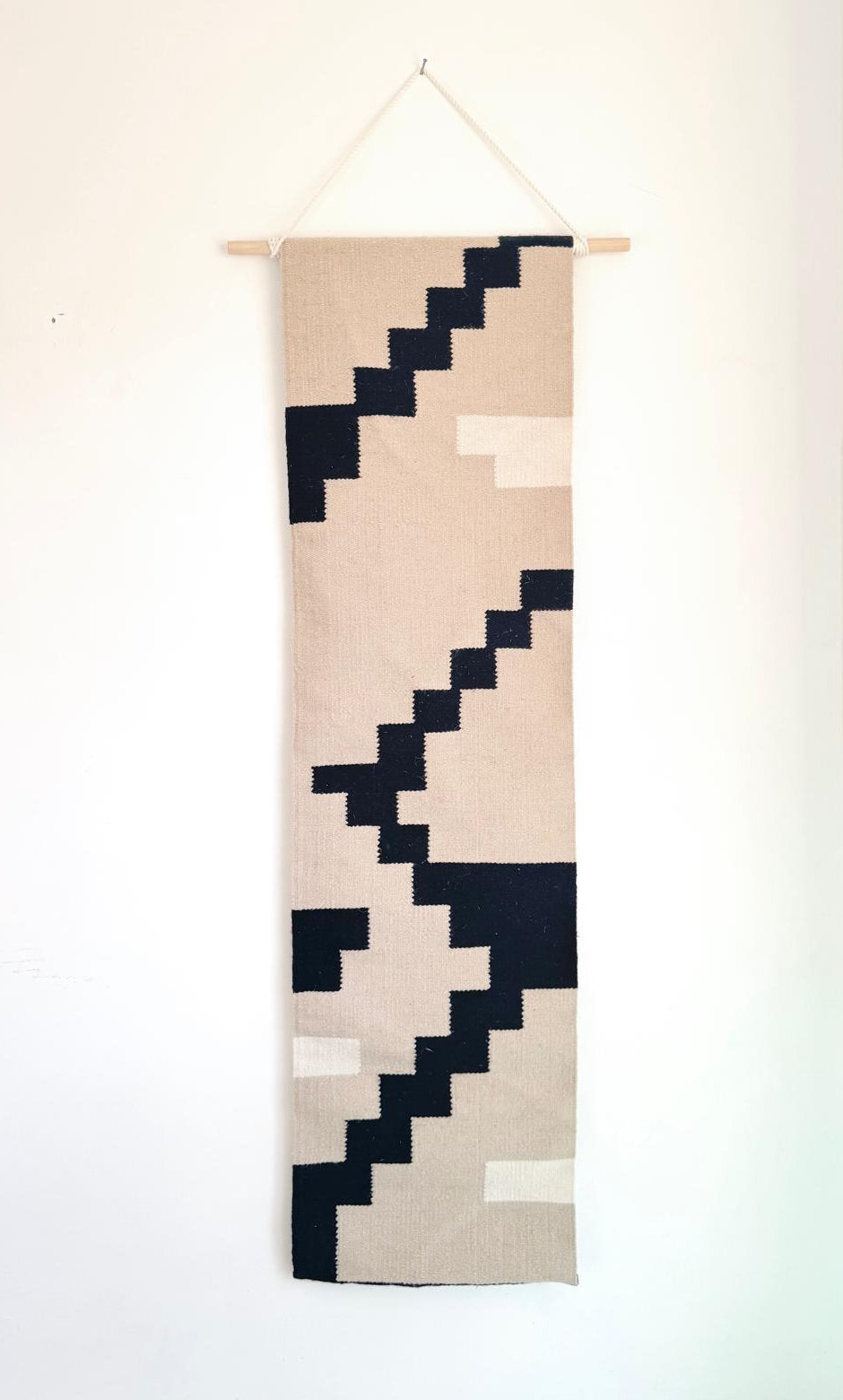 A vertical woven wall hanging tapestry with a geometric pixelated pattern in beige, cream, and black, suspended from a light wood dowel against a plain white wall.