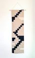 A vertical woven wall hanging tapestry with a geometric pixelated pattern in beige, cream, and black, suspended from a light wood dowel against a plain white wall.