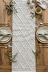 A woven off-white table runner with a raised diamond pattern, styled on a rustic wooden dining table with plates, cutlery, napkins, and olive branches.